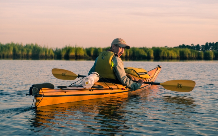 A young person sitting in a kayak looks back at the camera during a break from paddling. Trees line the shore in the background. 